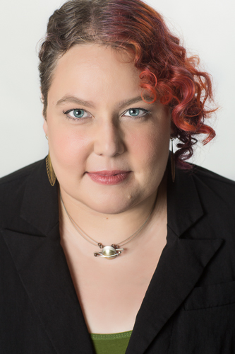 A professional headshot of Courtney. Courtney is wearing a back blazer, a green shirt, and a futuristic necklace. Their hair is curly with an asymmetrical cut and colored artificially red-orange on the long side. They're staring directly at the viewer with a slightly intense expression.